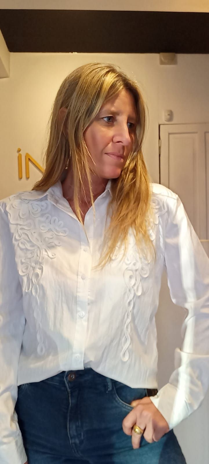 Mujer con blusa blanca y pantalones oscuros de pie en el interior de un edificio, mirando hacia un lado con una mano en el bolsillo.