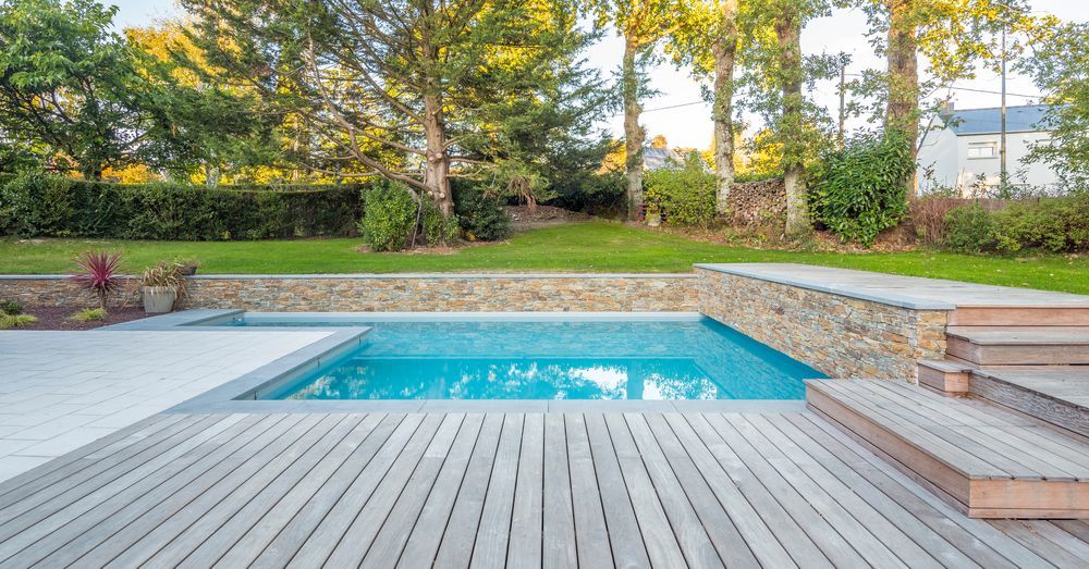 Swimming pool with wooden deck, stone wall, and green lawn. Turquoise water, sunny outdoor setting — Tru Blu Pools & Spas in Bluewater, QLD