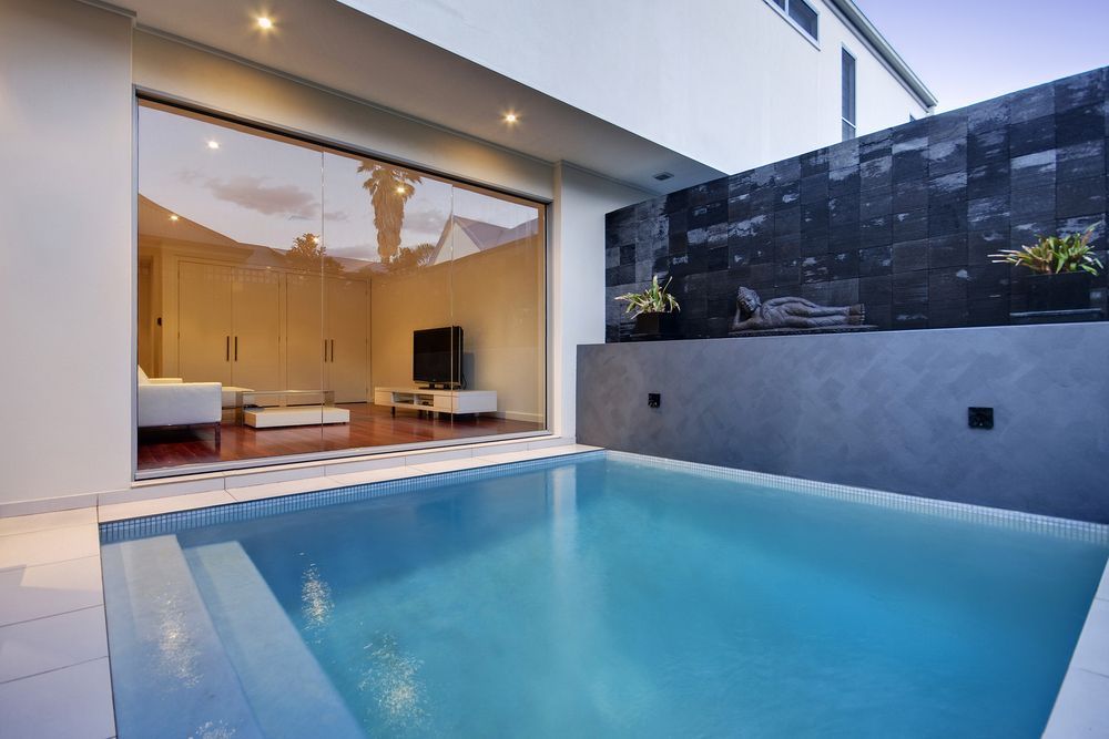 A small swimming pool outside a modern home, with glass sliding doors and a dark stone wall — Tru Blu Pools & Spas in Bluewater, QLD
