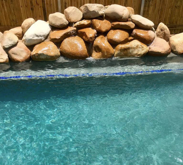 Pool with a rock waterfall. Clear blue water, beige and brown rocks, bamboo fence in background — Tru Blu Pools & Spas in Bluewater, QLD