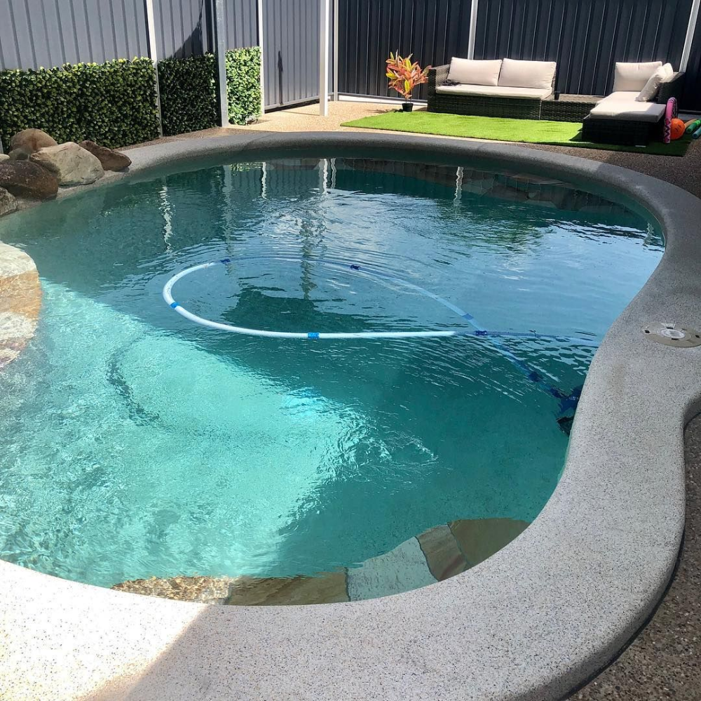 Oval-shaped swimming pool with blue water. A robotic pool cleaner is submerged. Surrounded by stone and greenery— Tru Blu Pools & Spas in Bluewater, QLD