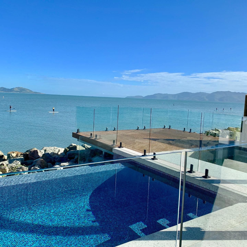 Infinity pool overlooking ocean, with a wooden deck and two paddle boarders in the distance under a blue sky— Tru Blu Pools & Spas in Bluewater, QLD