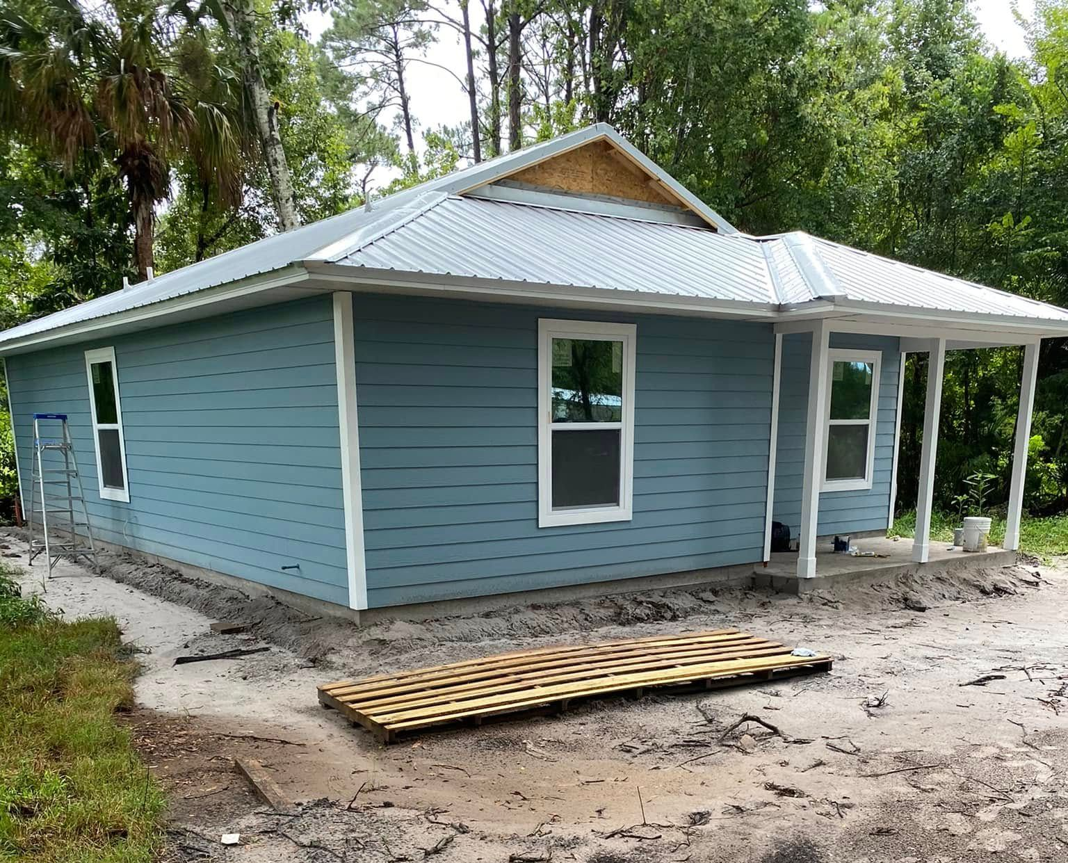 Single-story blue house with white trim and porch, surrounded by trees and unfinished dirt yard