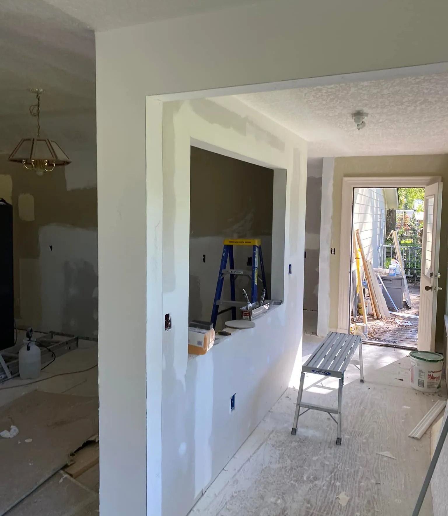 Renovation hallway with drywall, ladder, bench, and doorway opening to a bright room and outdoor patio.