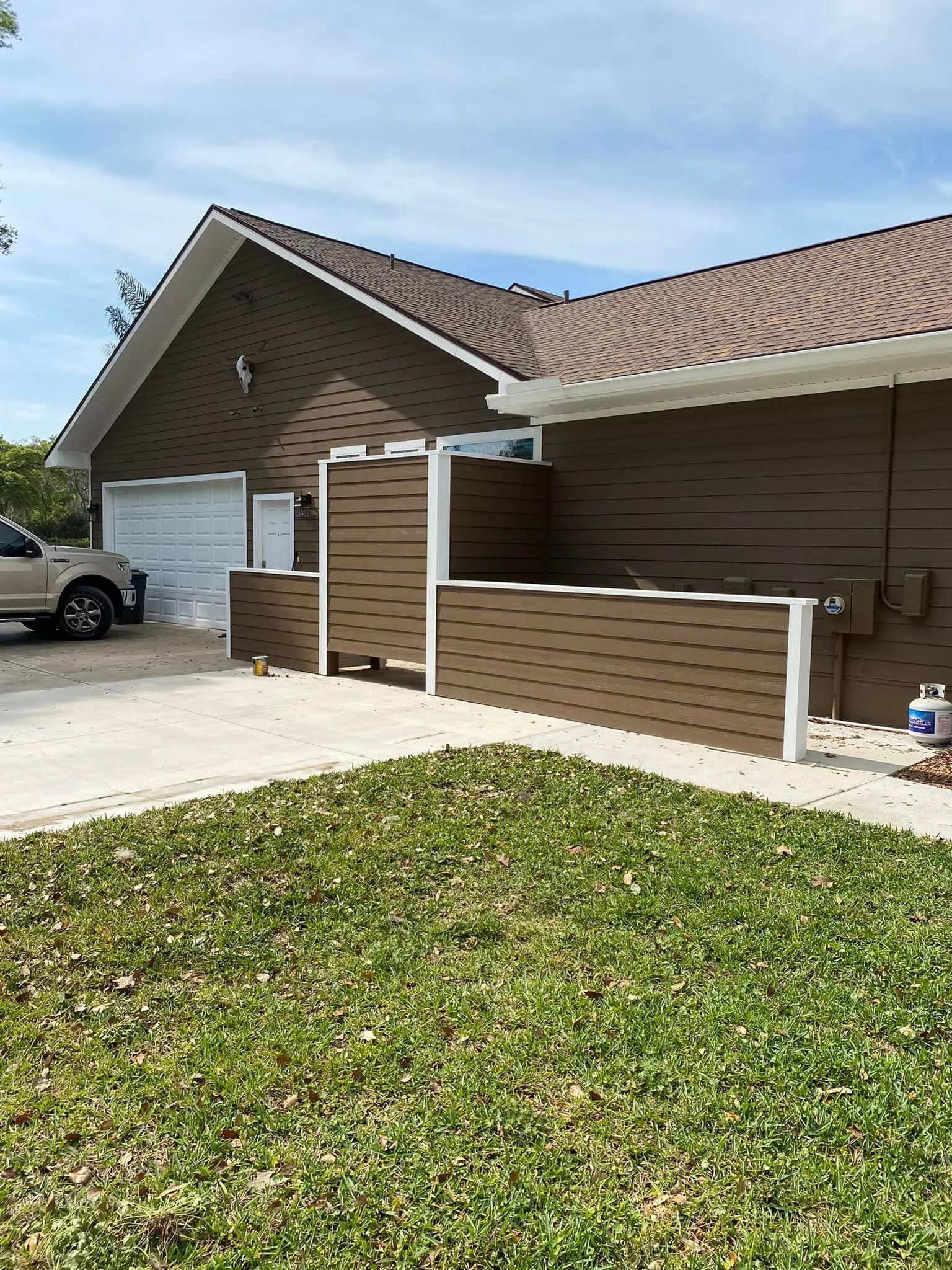 Suburban house with brown siding, white trim, a garage, and a small fenced patio under a blue sky