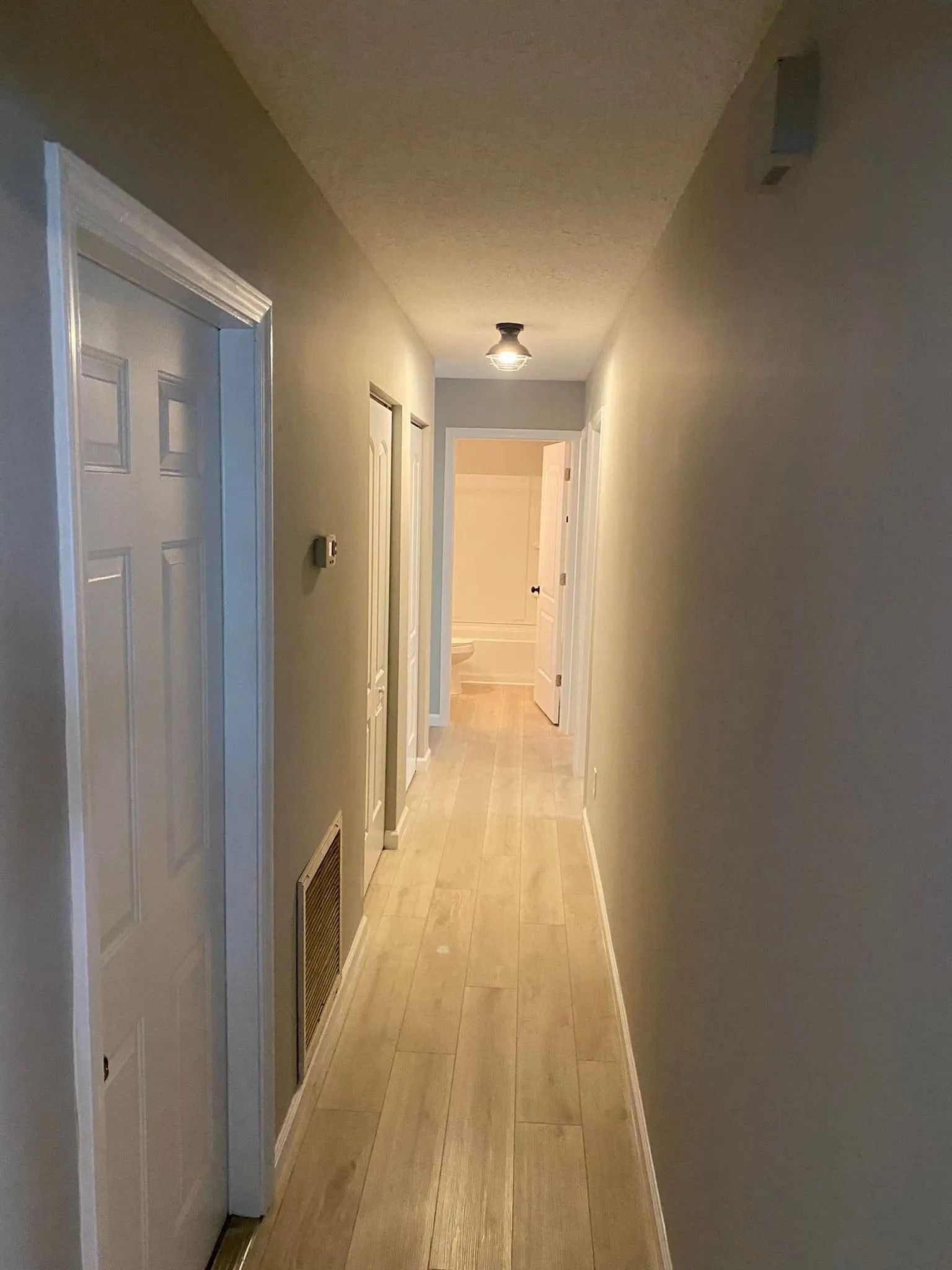 Long, narrow hallway with white walls, wood floor, doors, vents, and a ceiling light at the end.