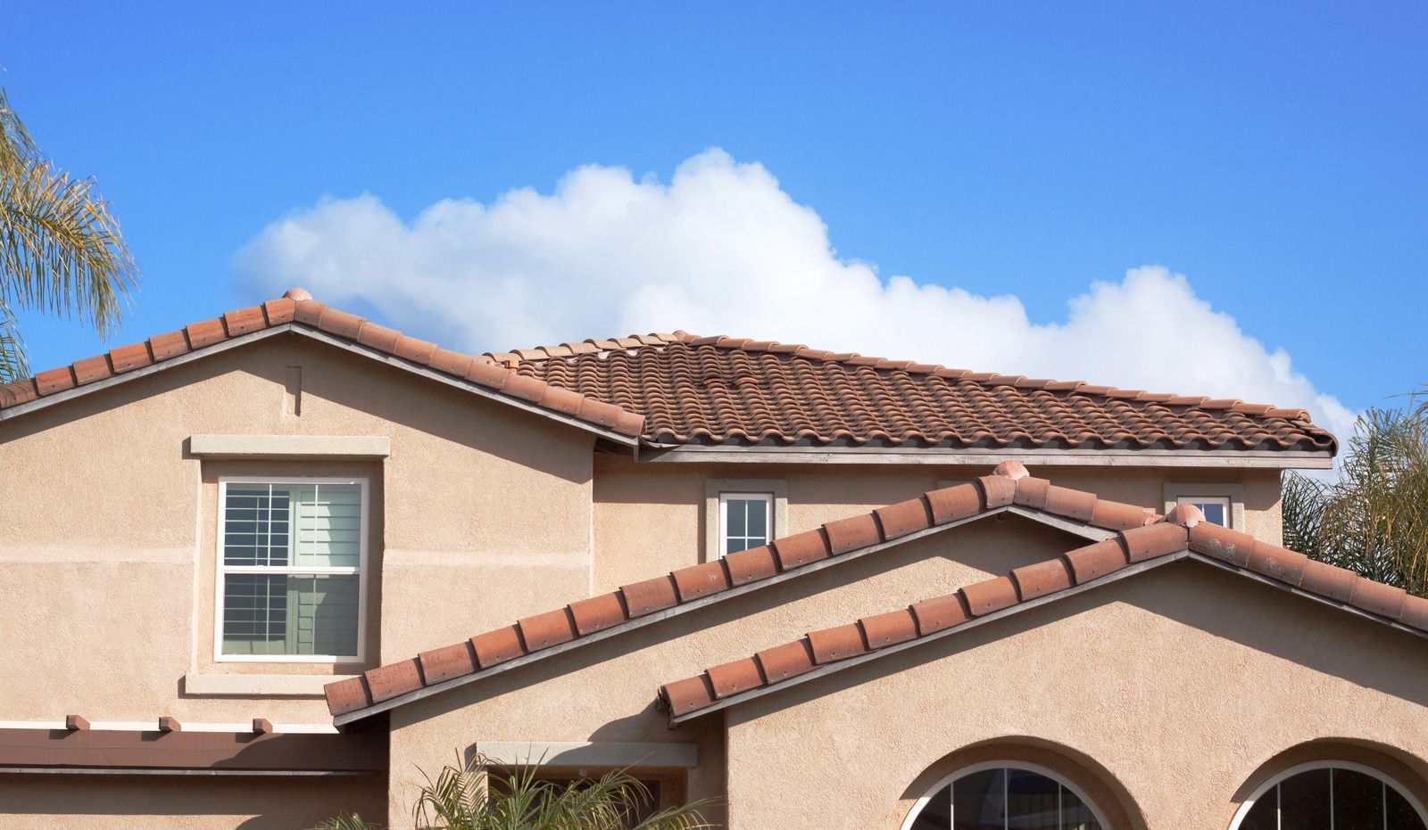 Two-story beige house with red tile roof against a blue sky