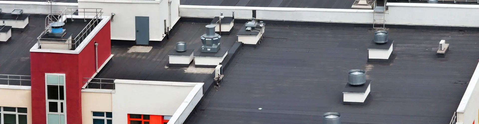 View of a flat, dark roof with various ventilation units, surrounded by white structures. A red building is on the left.