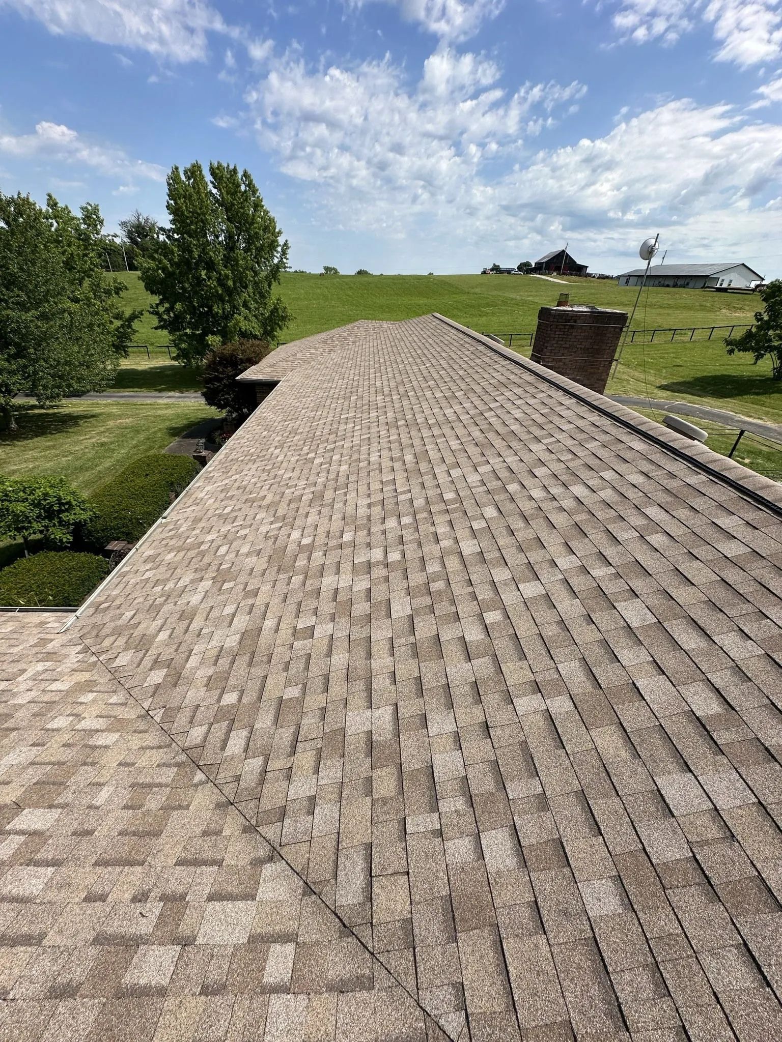 View from a brown shingle roof on a sunny day, overlooking green fields and trees.