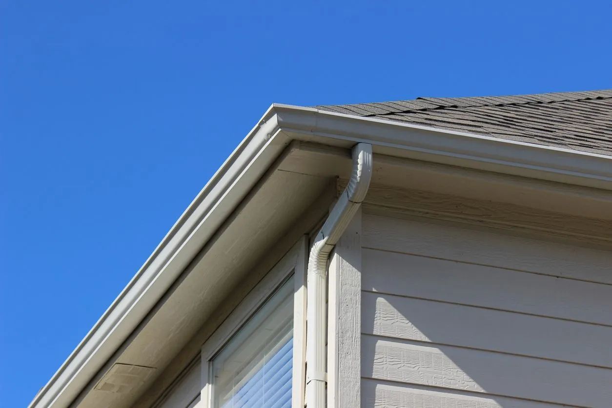 Beige house corner with gray roof, gutters, and downspout against a bright blue sky.