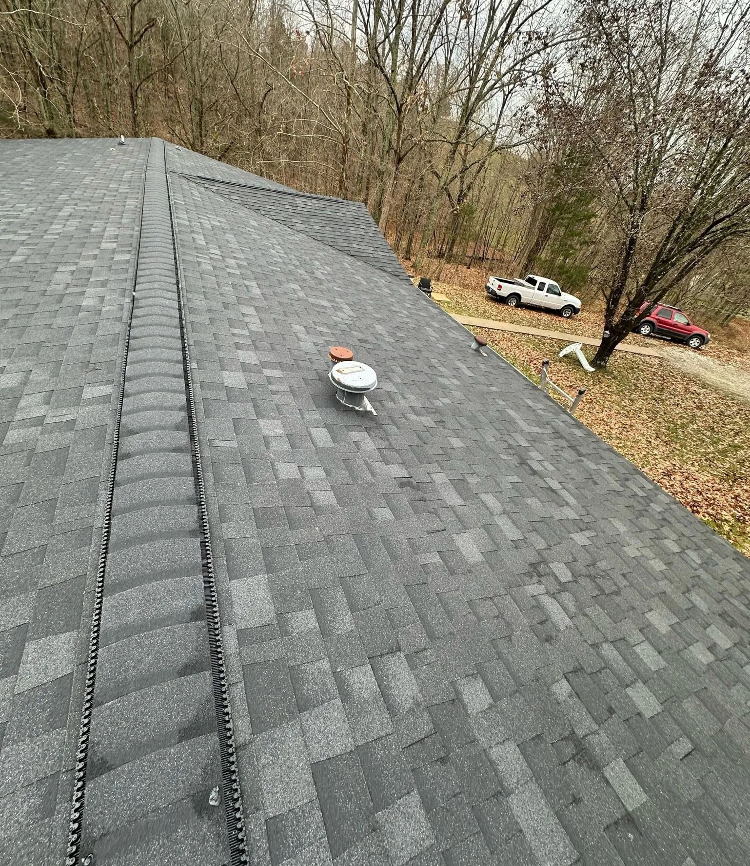 Gray shingle roof with a vent pipe, viewed from above near bare trees and parked vehicles below