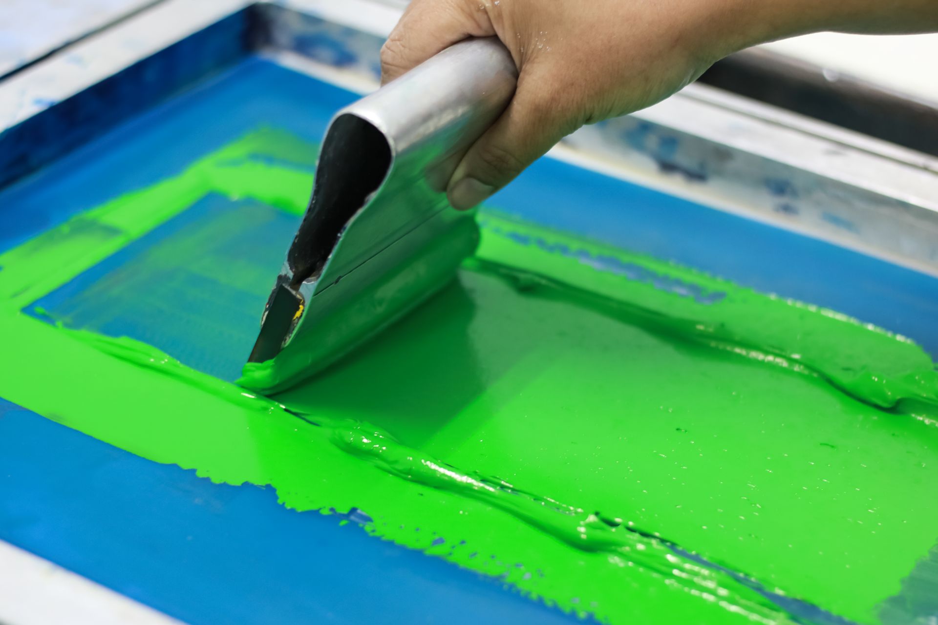 A hand uses a squeegee to spread bright green ink across a blue screen printing frame. A hand uses a squeegee to spread bright green ink across a blue screen printing frame.