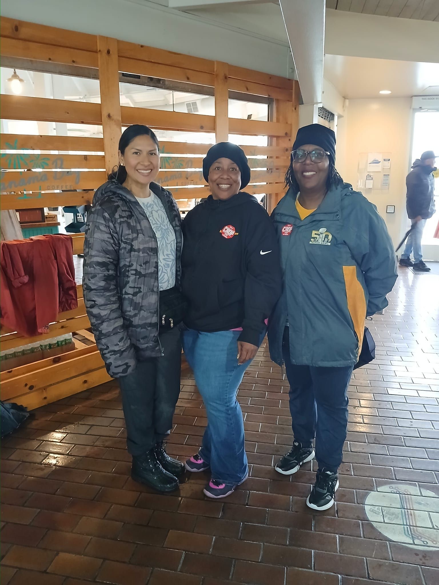 Three women stand indoors. One wears a camo jacket, one a black hoodie, and the third a gray jacket.