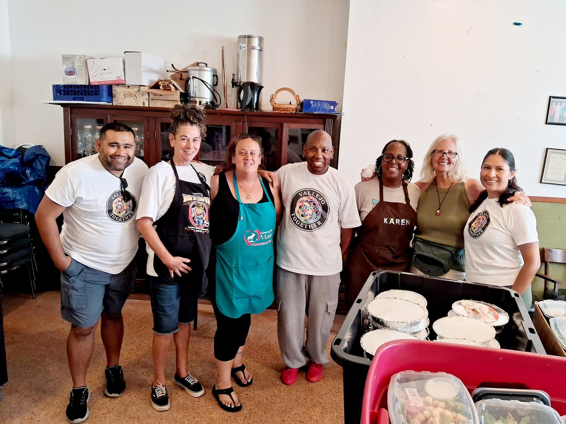 Group of people wearing aprons, smiling, posing near prepared food. Interior shot.
