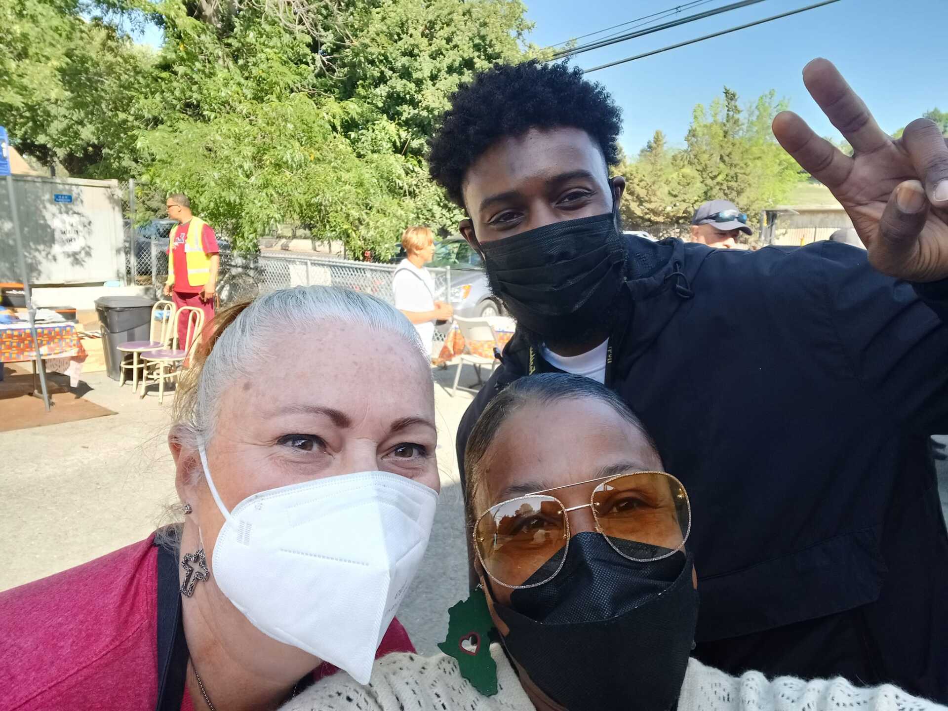 Three people wearing masks pose for a selfie outdoors. Two smile, and one gives a peace sign. Bright sunlight.