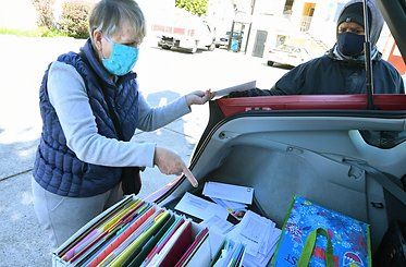 Two people in masks sorting papers from a car trunk outdoors.