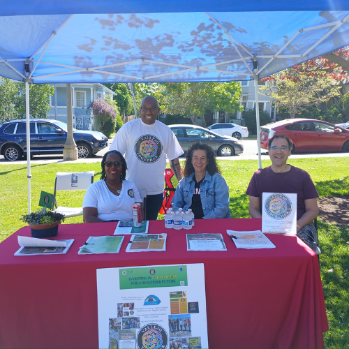 People at a table under a blue canopy; brochures and information are displayed, outside on a sunny day.