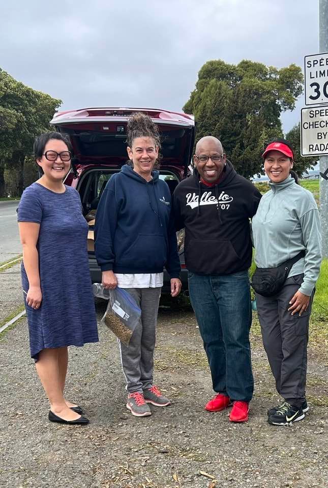 Four people standing near a vehicle; two women and two men. Cloudy, outdoor setting with a speed limit sign.