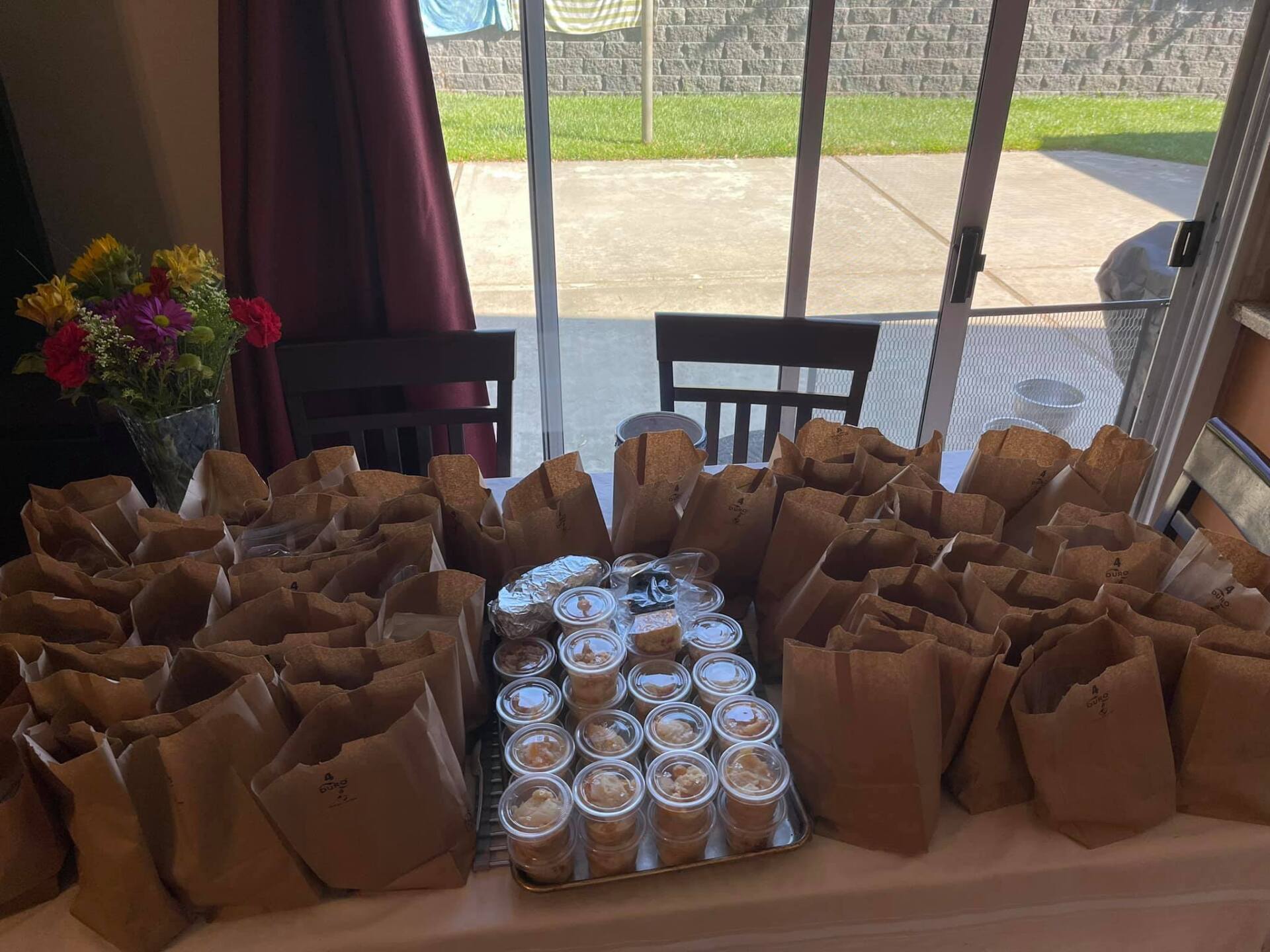 Table set with many brown paper bags and containers of food, flowers in a vase behind them.