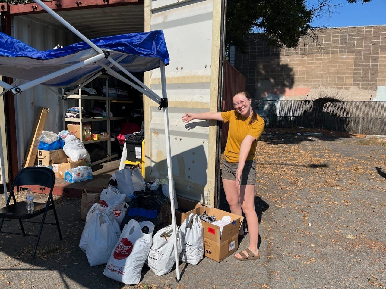 Woman points at donated supplies in front of a blue-canopy tent and building.