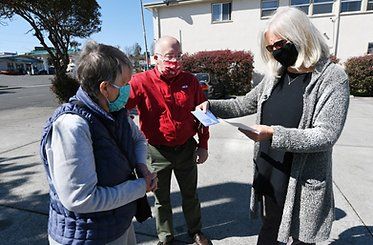 Three people wearing masks stand outside. Woman shows paper to two people, the other two look on.