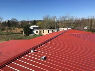 A bright red metal roof on a sunny day, with a glimpse of a nearby building and bare trees in the background.