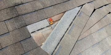 A damaged roof with several missing shingles, revealing the wooden roof deck and a small orange marker underneath.