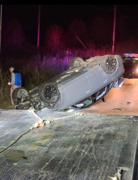 Car overturned on a tow truck, illuminated by emergency lights. A person stands nearby. Nighttime setting.