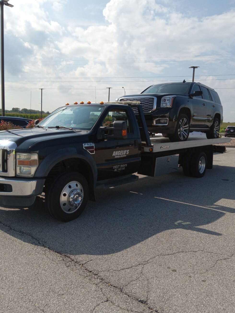 Black tow truck carrying a black SUV on a flatbed. Sunny outdoor setting.