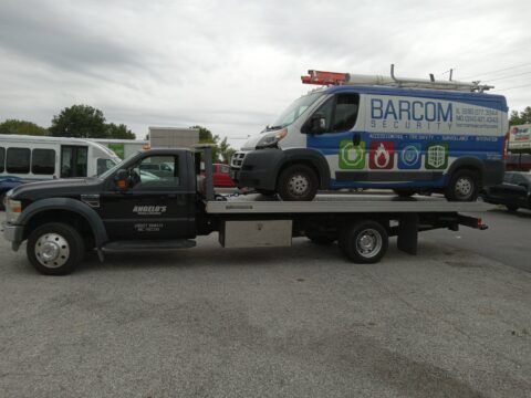 Black tow truck carrying a white and blue BARCOM SECURITY van on its flatbed in a parking lot.