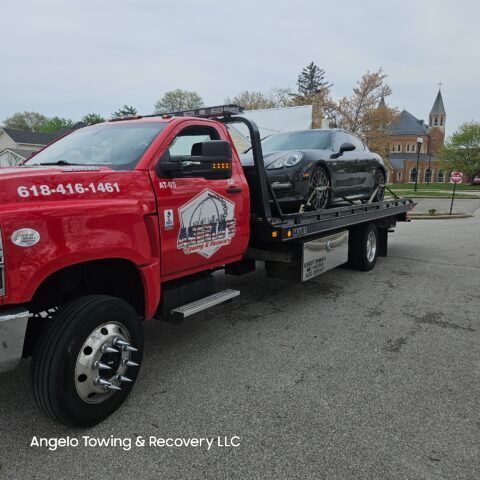 Red tow truck hauling a black sports car in front of a church.