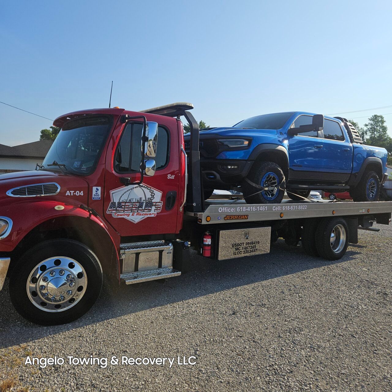 Red tow truck towing a blue pickup truck on a gravel lot under a clear sky.