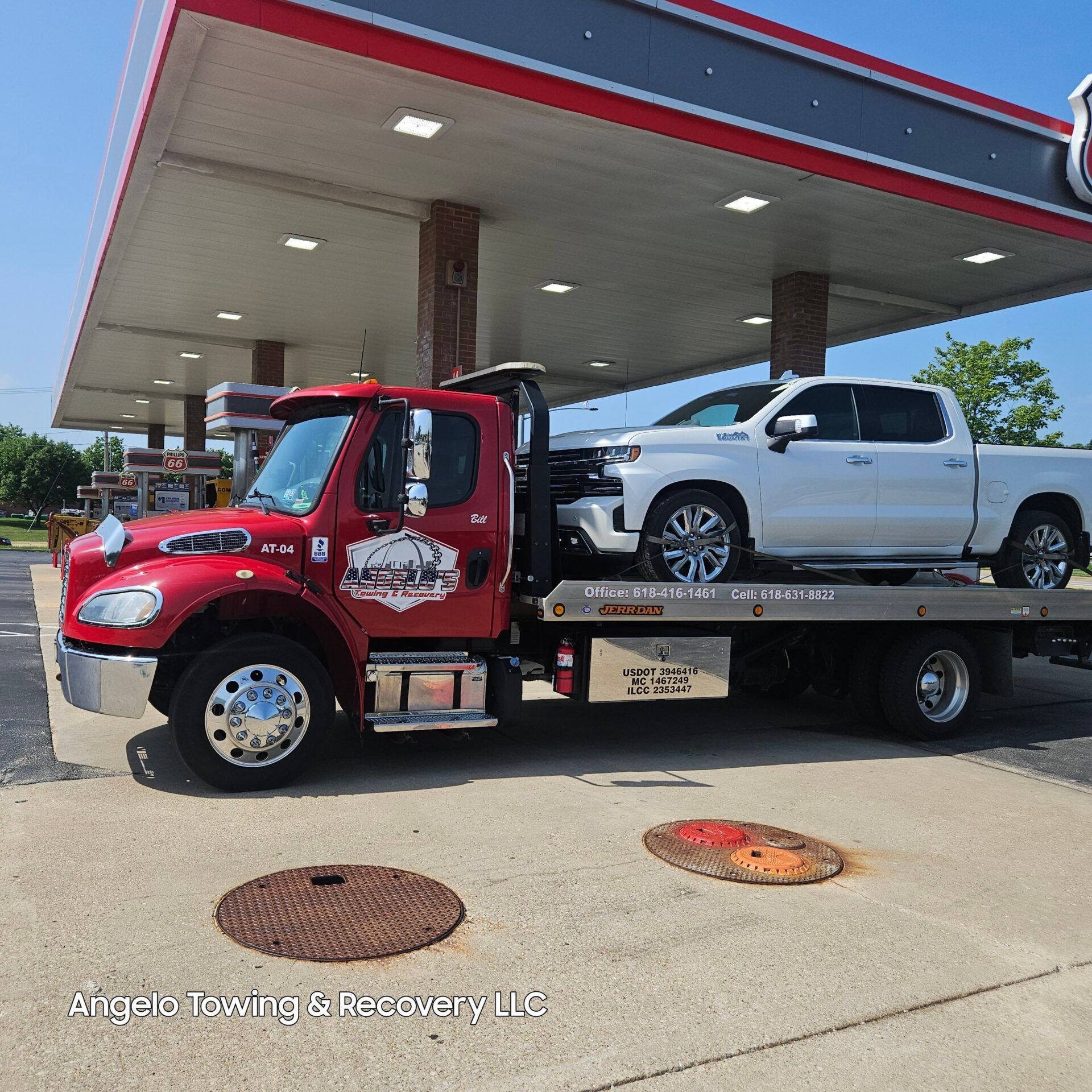 Red tow truck with a white pickup truck on its bed at a gas station.