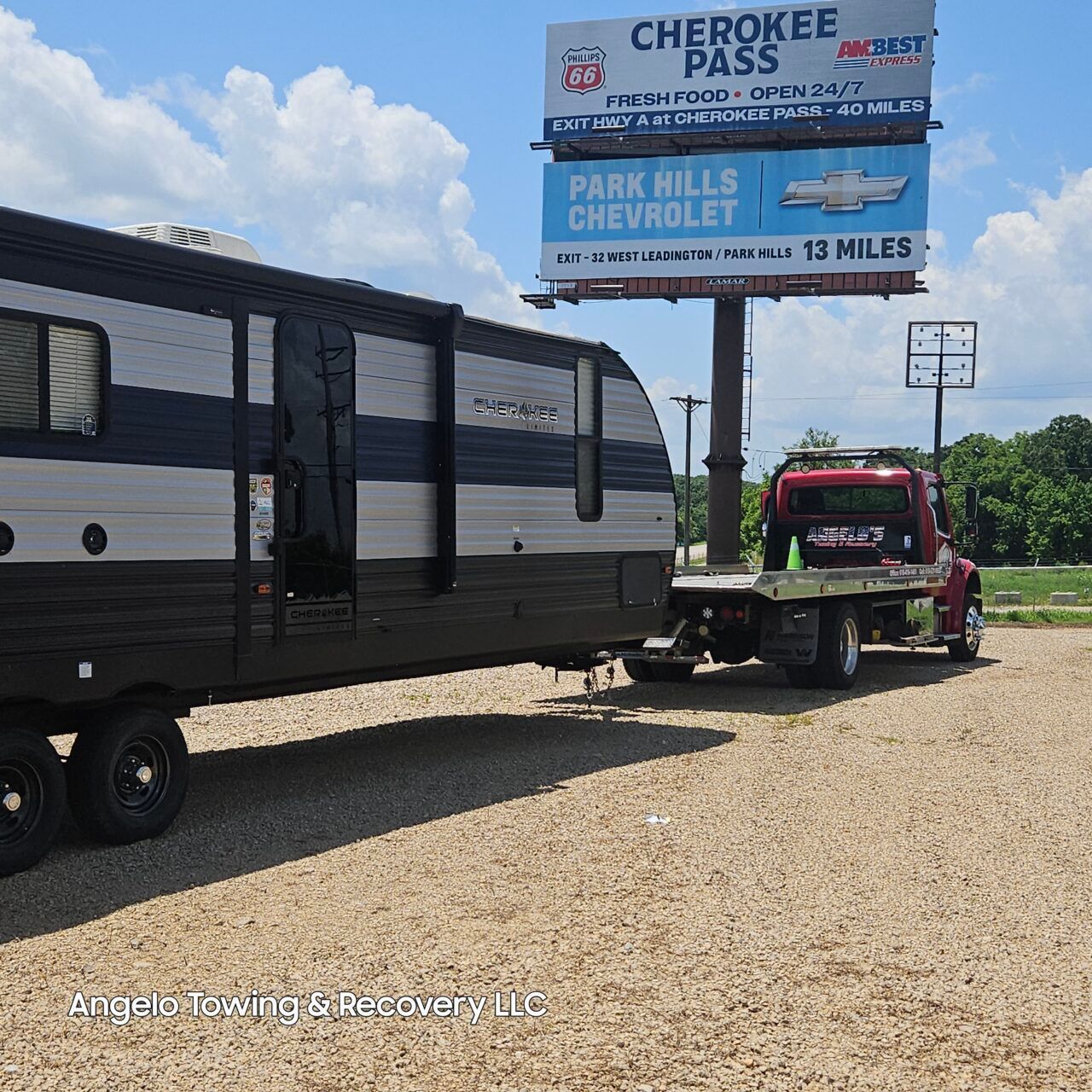 A red tow truck towing a large travel trailer on gravel, with a billboard in the background.