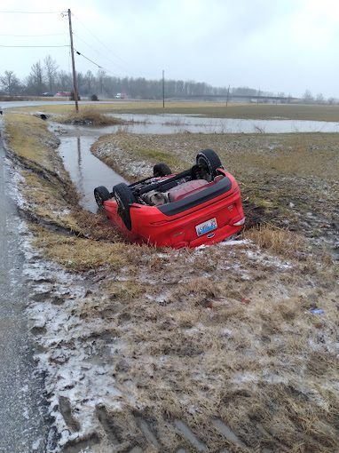 Red car overturned in a flooded ditch next to a road, in a rural setting.
