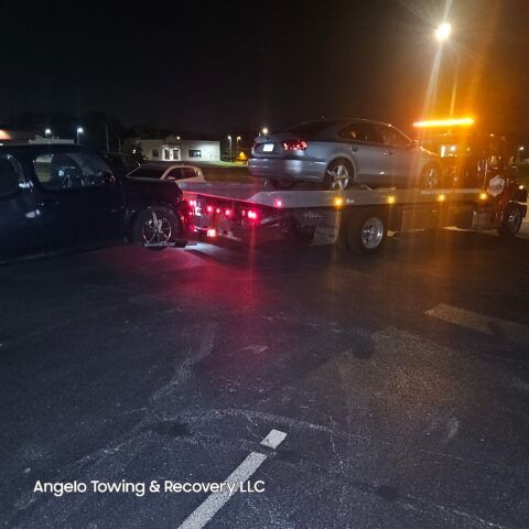 A tow truck loading a silver car in a dark parking lot at night. Orange lights illuminate the scene.