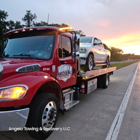 Red tow truck with a white pickup truck on its flatbed, sunset in the background.