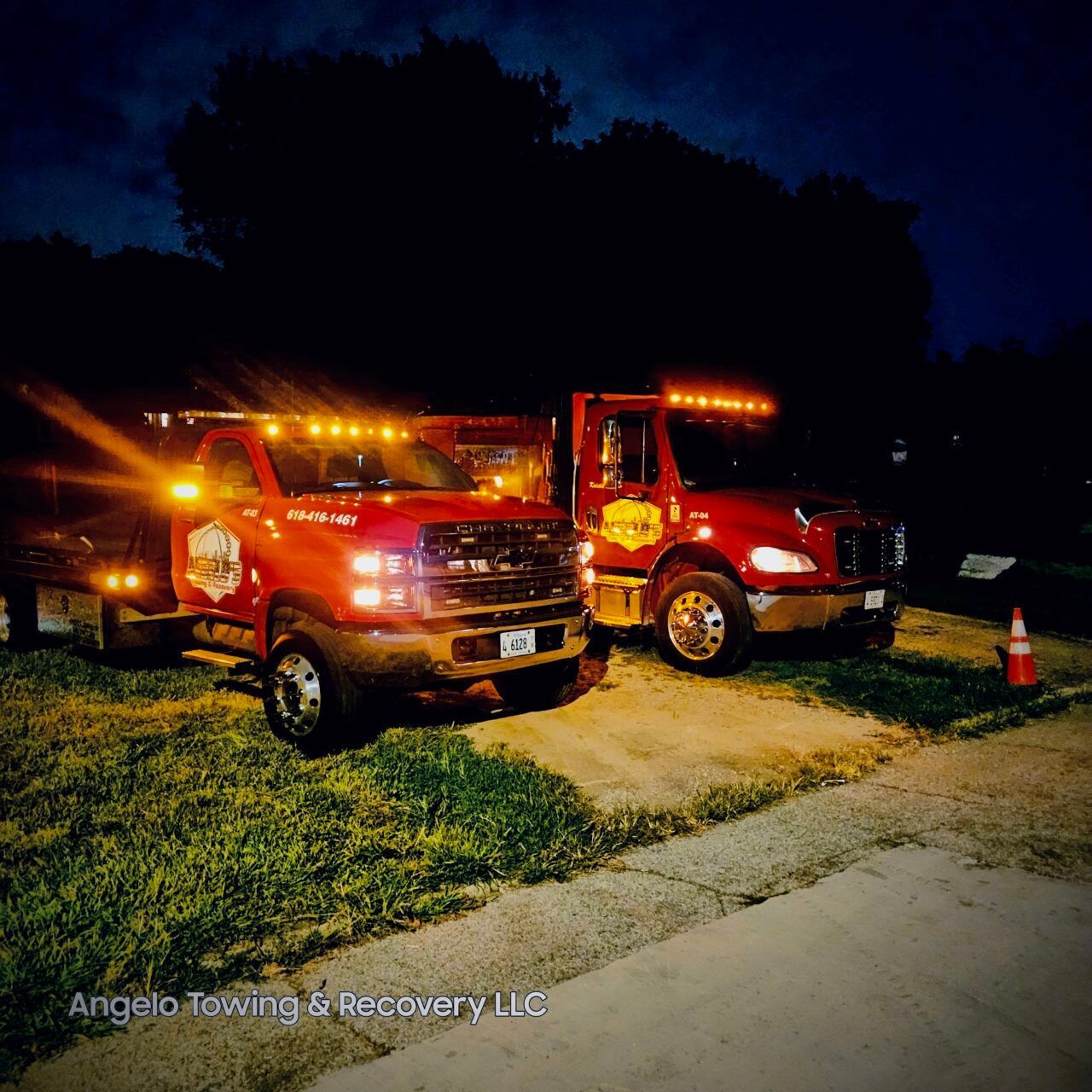 Two red tow trucks parked side-by-side at night, lights on. Grass and a dark sky in the background.
