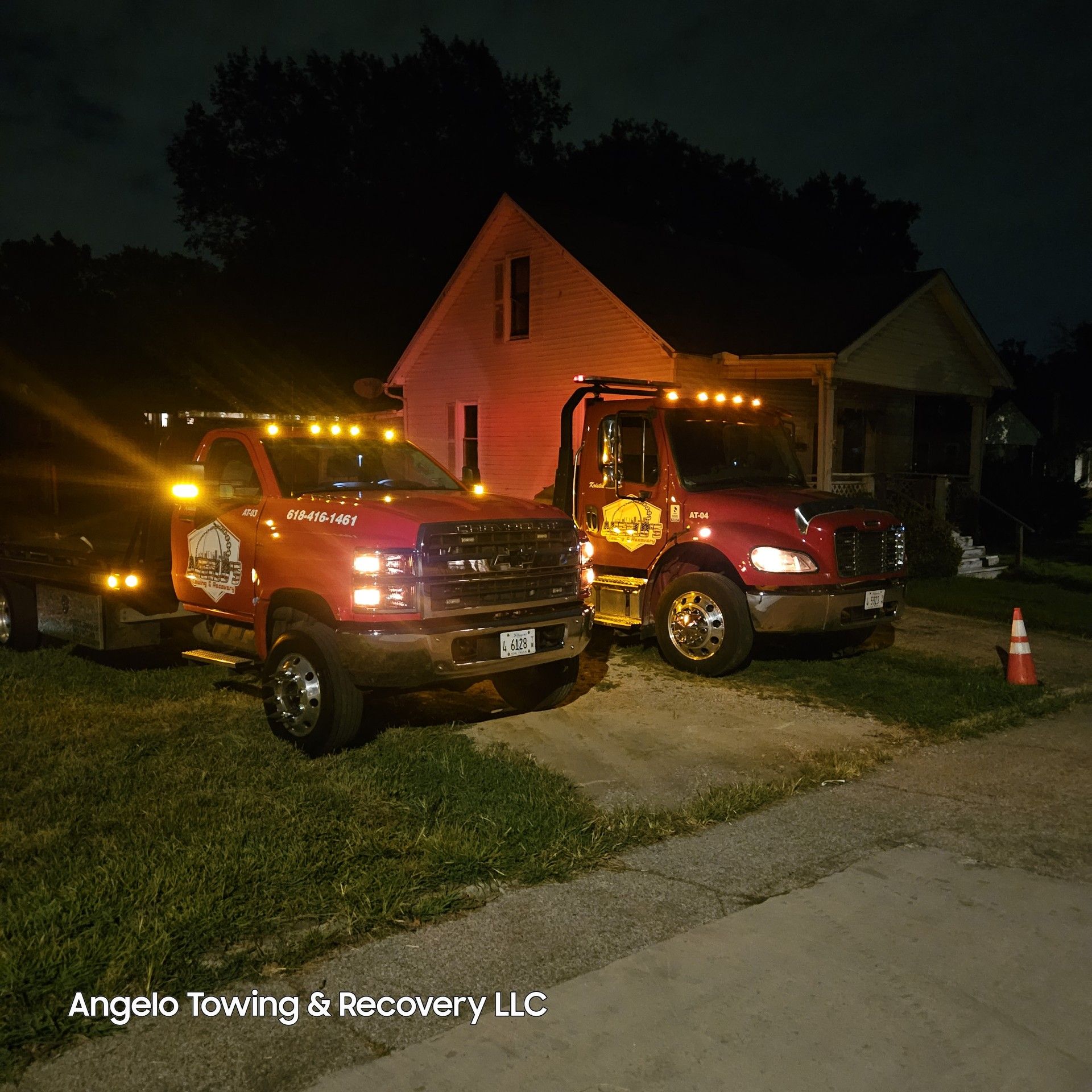 Two Angelo Towing & Recovery trucks parked at night in front of a house, lights on.