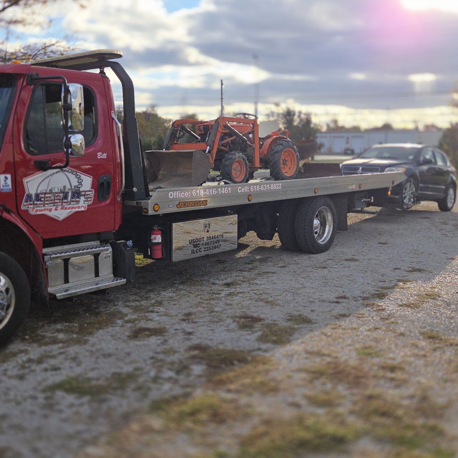 Red tow truck carrying a small orange tractor and a black car on a sunny day.