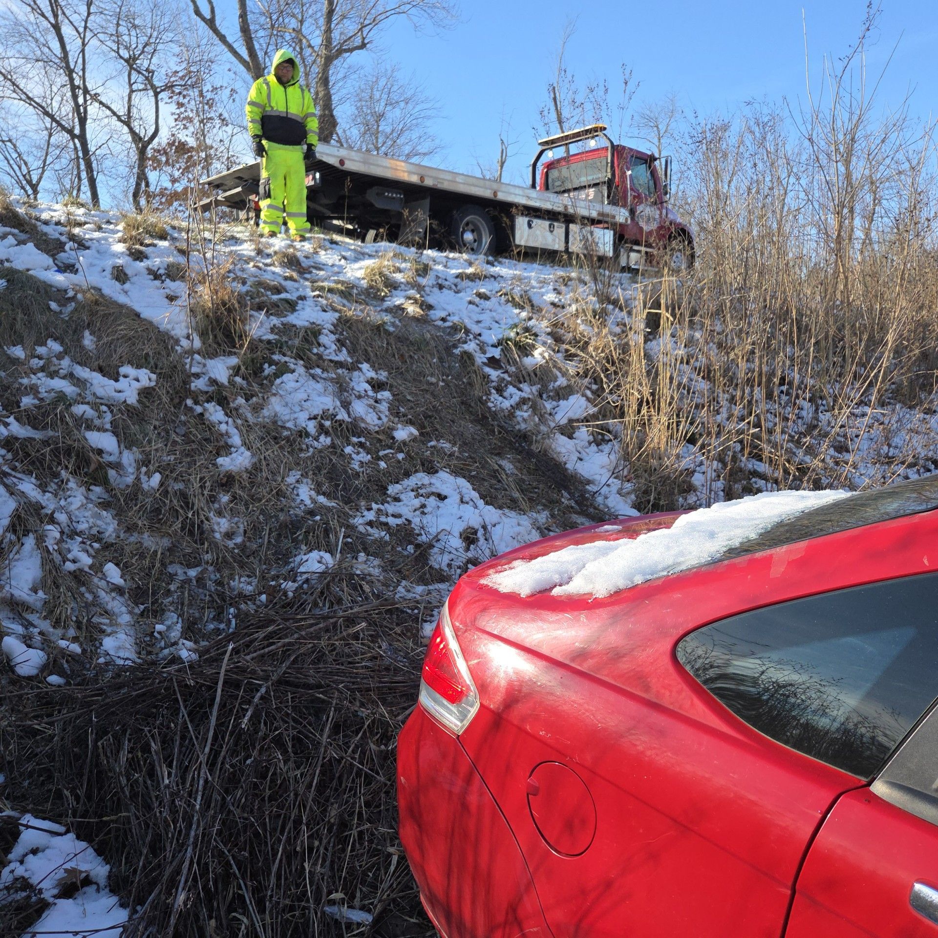 Red car in a ditch being retrieved by a tow truck; tow truck driver in a yellow suit stands by. Snowy scene.