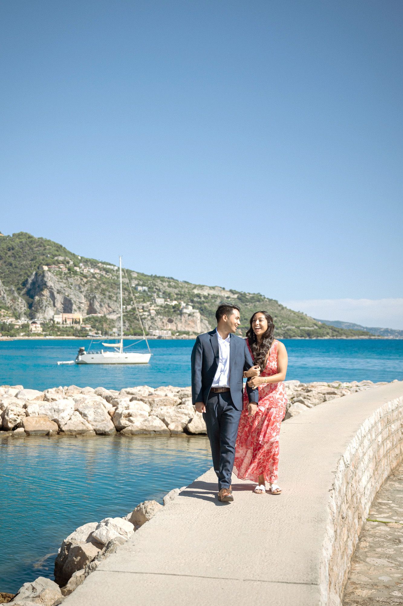 A couple laughing and smiling during a photoshoot with the sea and mountains in the background.
