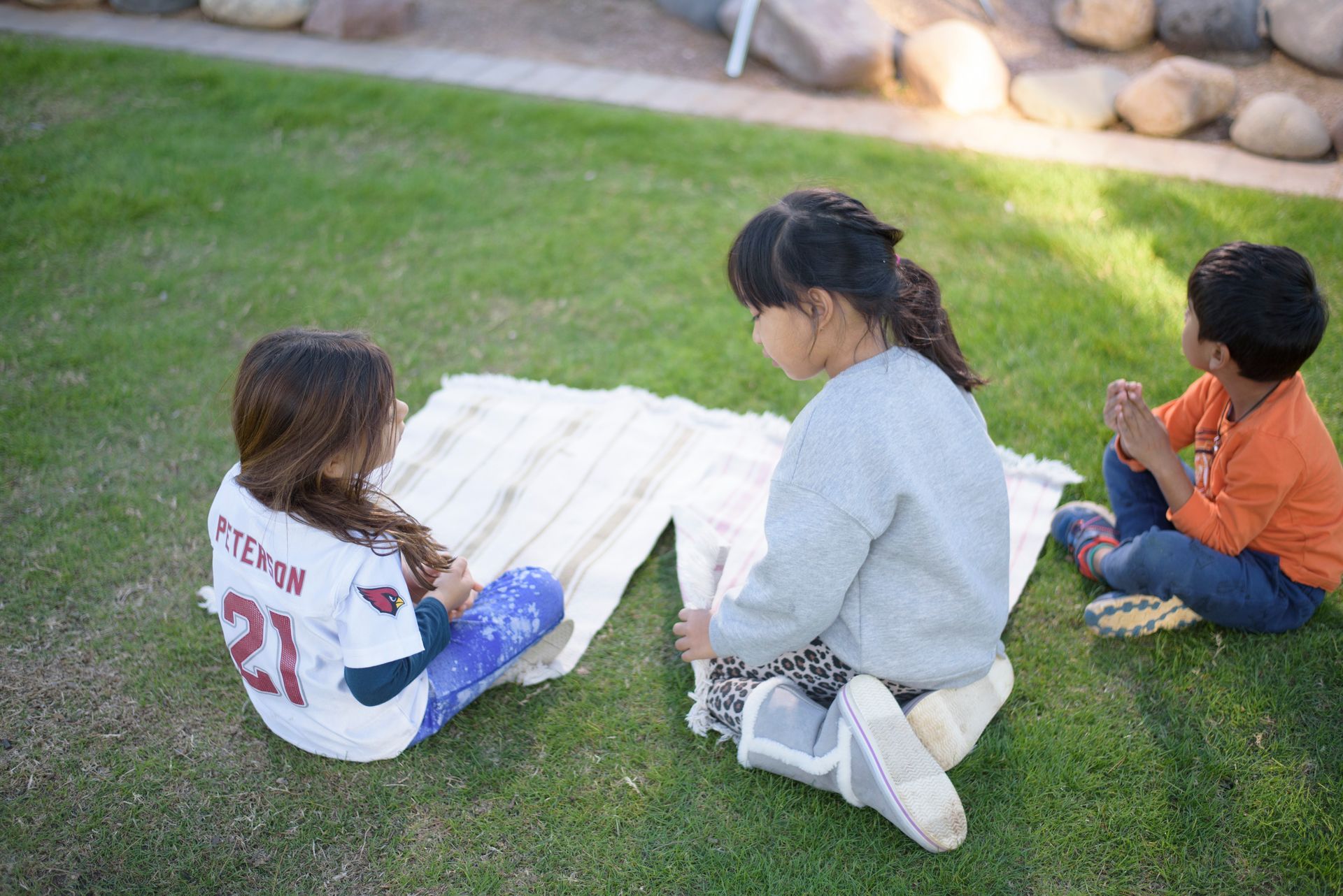 Three children sit on grass around a white picnic mat, facing away outdoors.