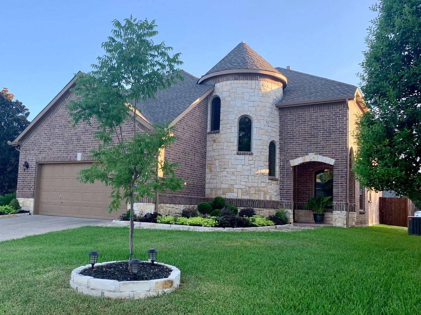 Stone suburban house with turret, arched entry, and two-car garage, fronted by a green lawn and young tree