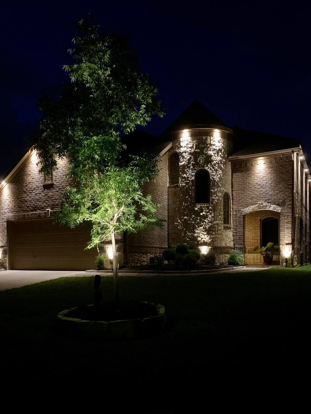Well-lit stone house at night with a tree in the front yard
