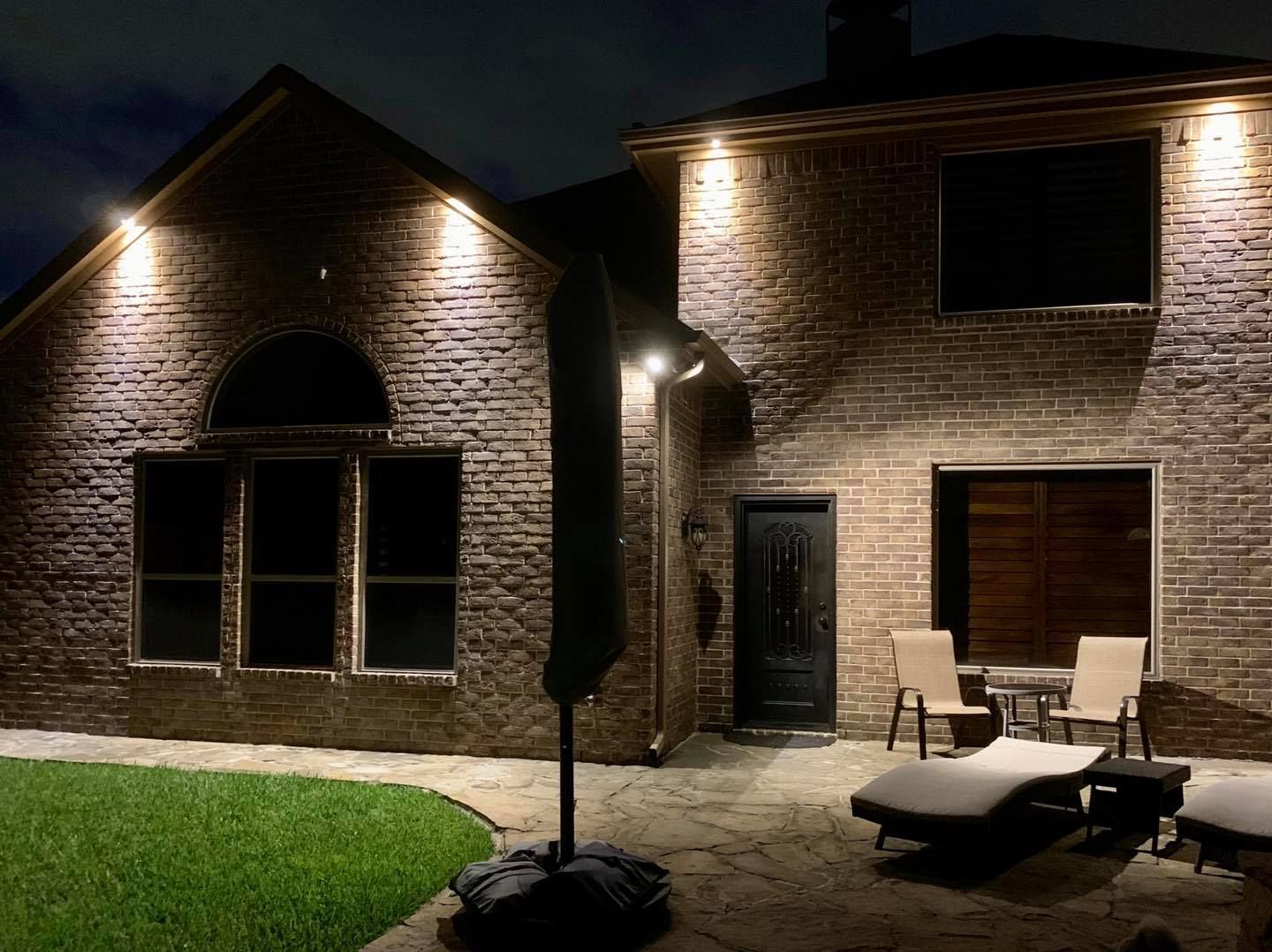 Brick house patio lit at night, with chairs, lounge chair, and a dark lawn.