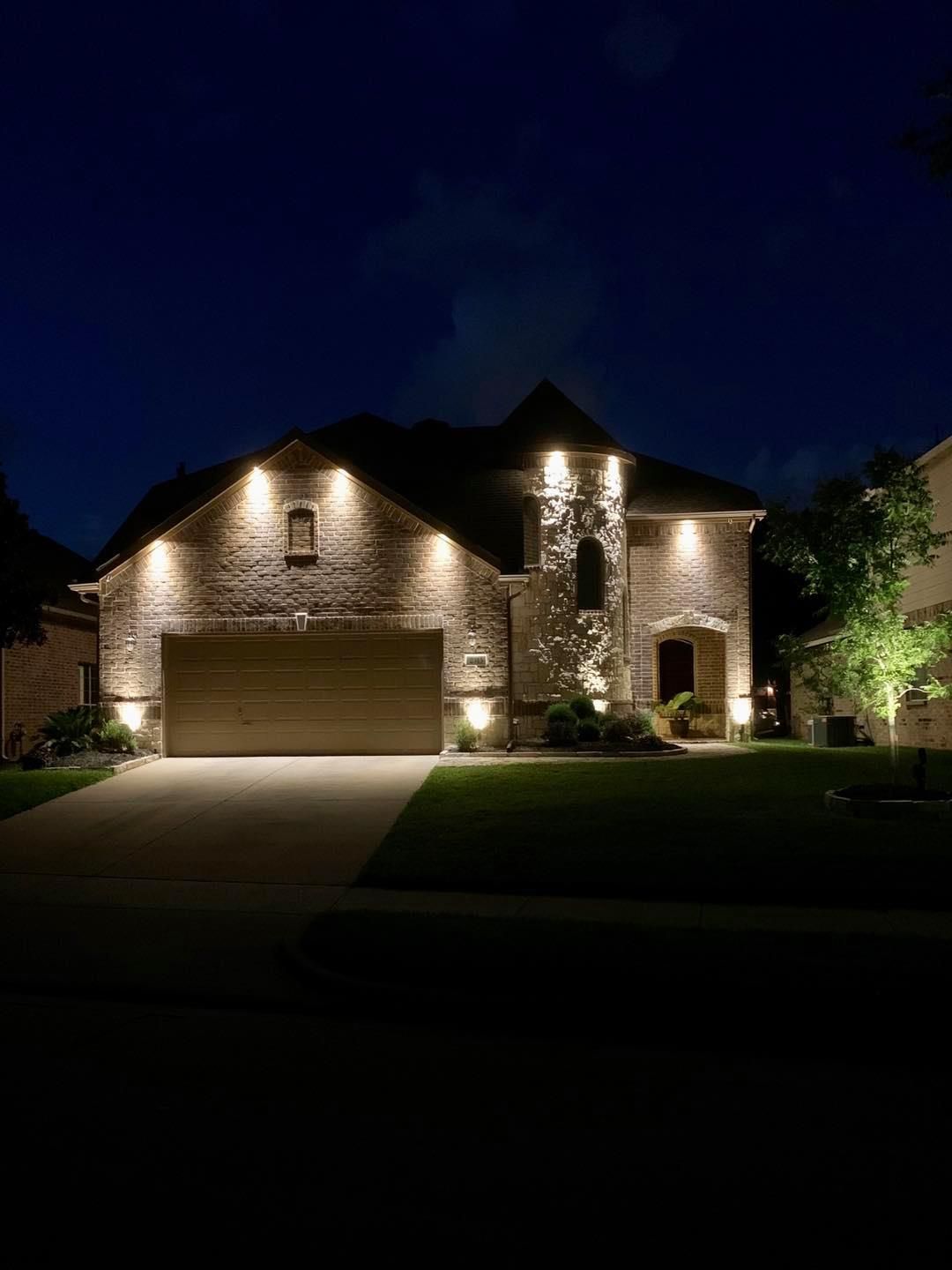Nighttime view of a stone house with warm exterior lights and a lit garage, framed by a dark sky and lawn