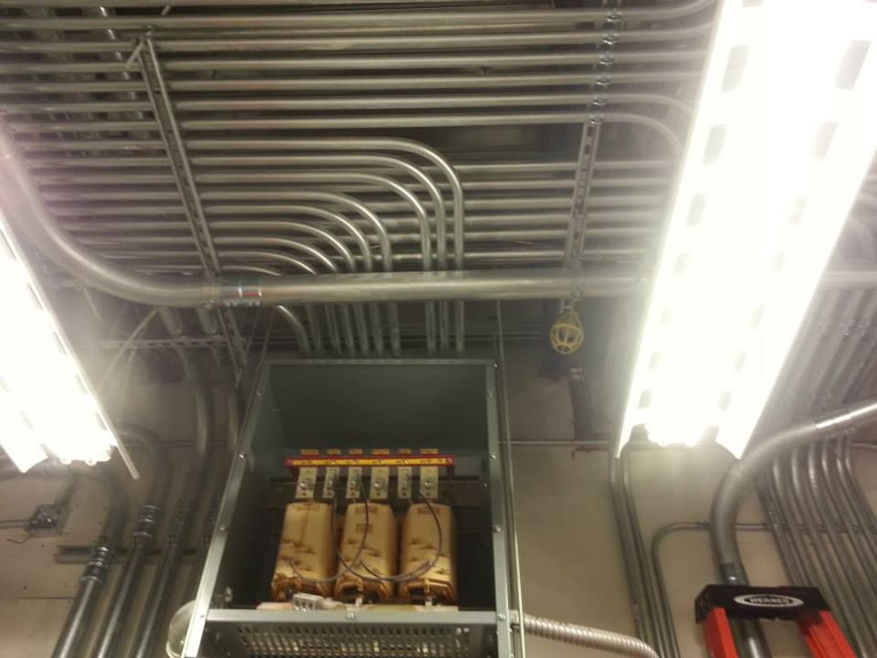 Electrical panel with exposed breakers and wiring beneath bright fluorescent ceiling lights