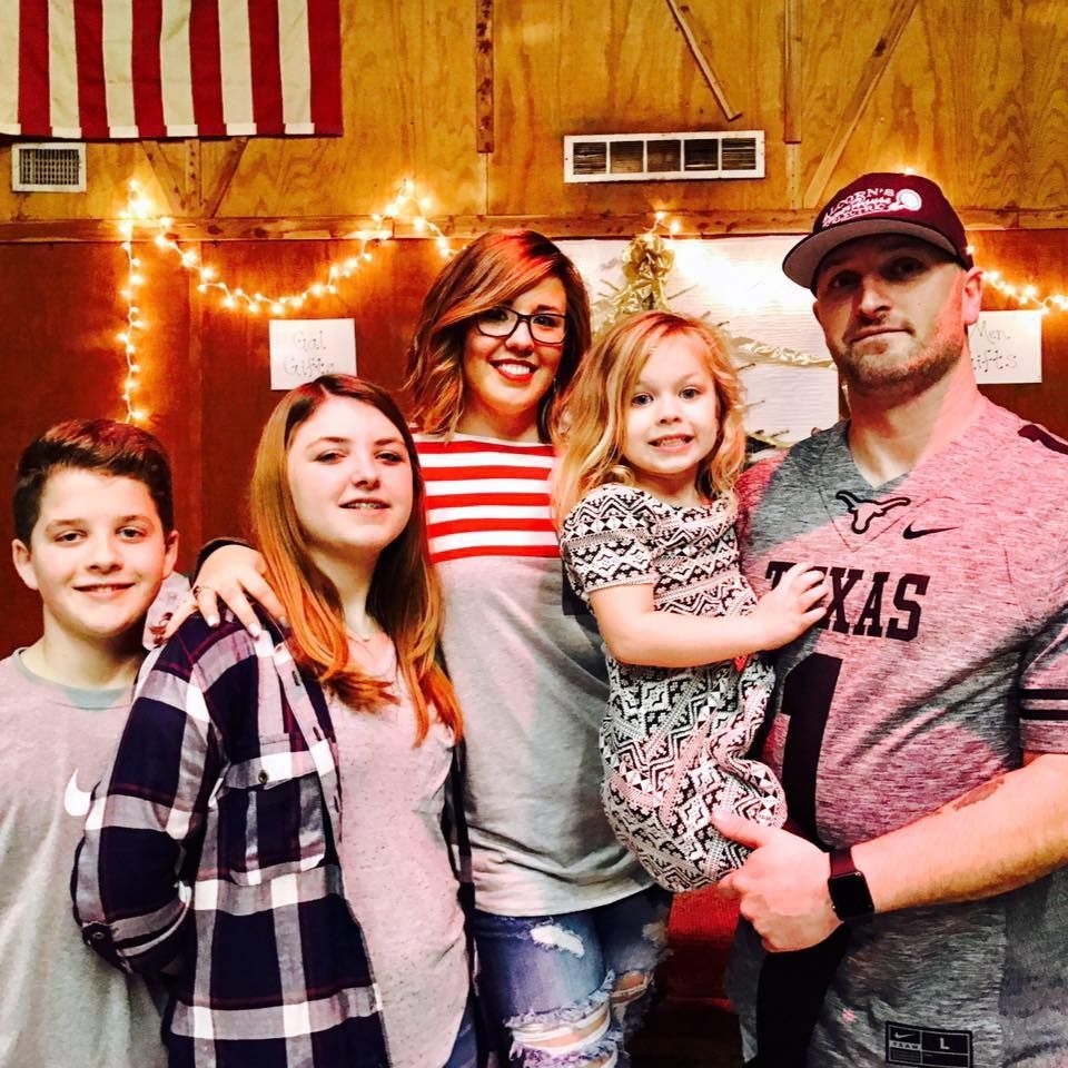Five people posing indoors; a man holds a young girl in front of warm string lights.