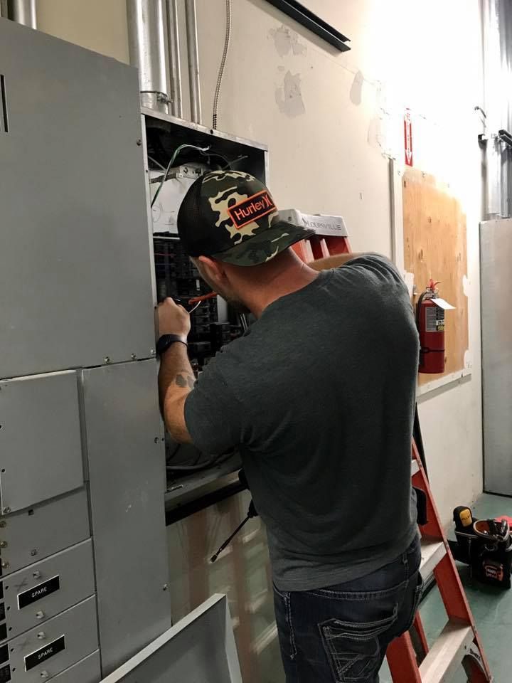 Worker servicing electrical panel in a utility room with exposed wiring.